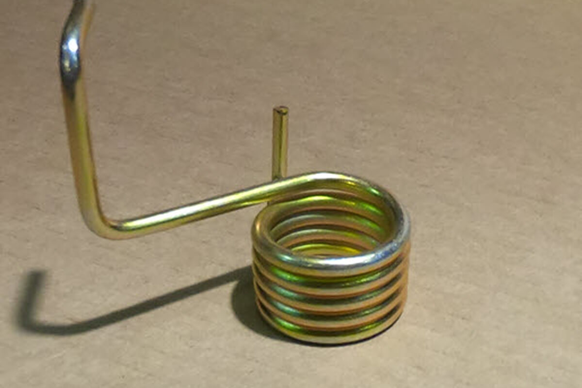 Close-up of a metallic torsion spring with a coiled body and extended arms, placed on a cardboard surface.